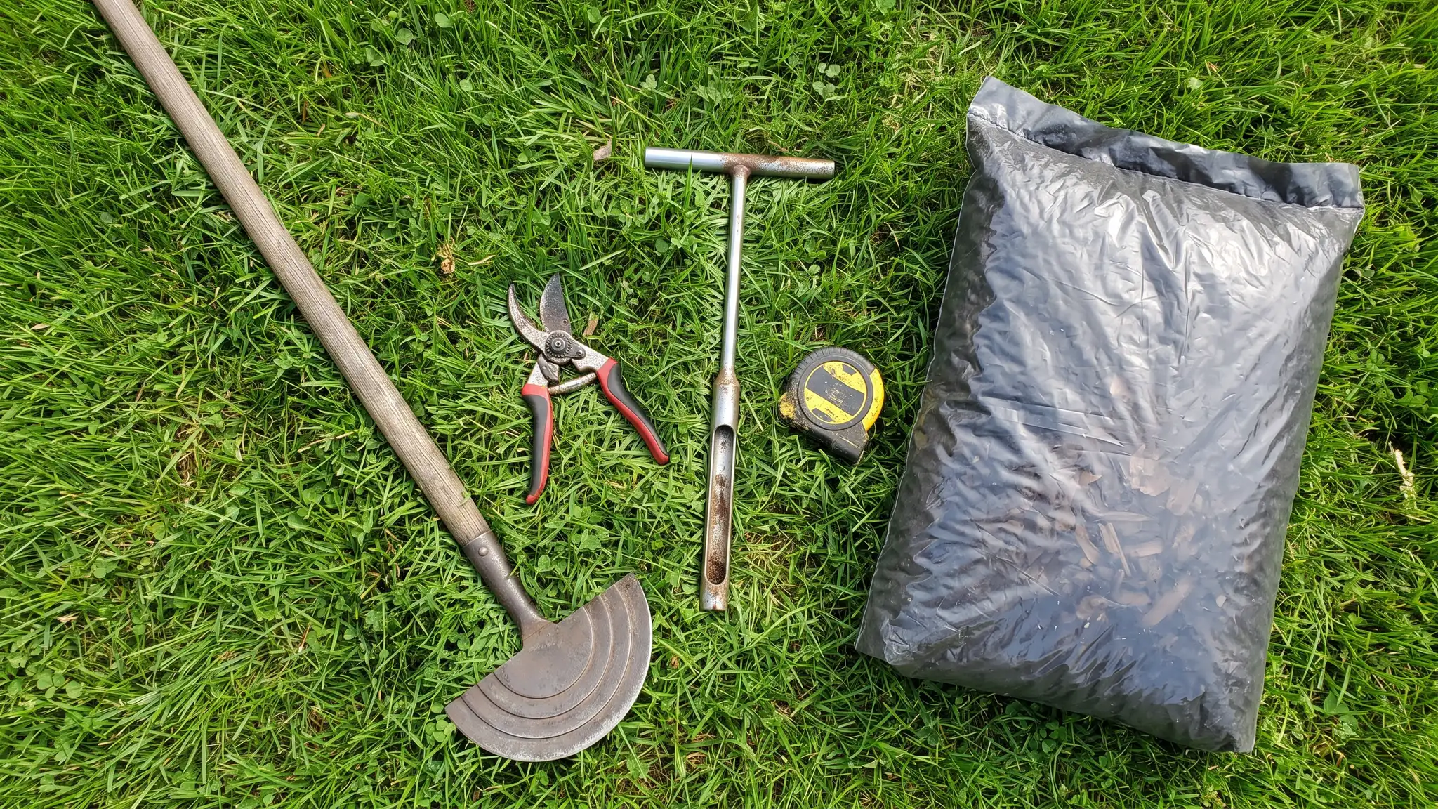 A flat lay on green grass of lawn care tools: a hand edger, pruning shears, a soil probe and a tape measure